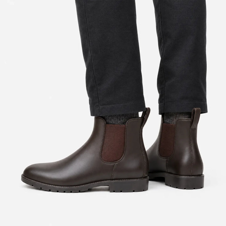 Side view of brown Chelsea rain boots worn with dark charcoal pants and gray socks against a plain white background. 4