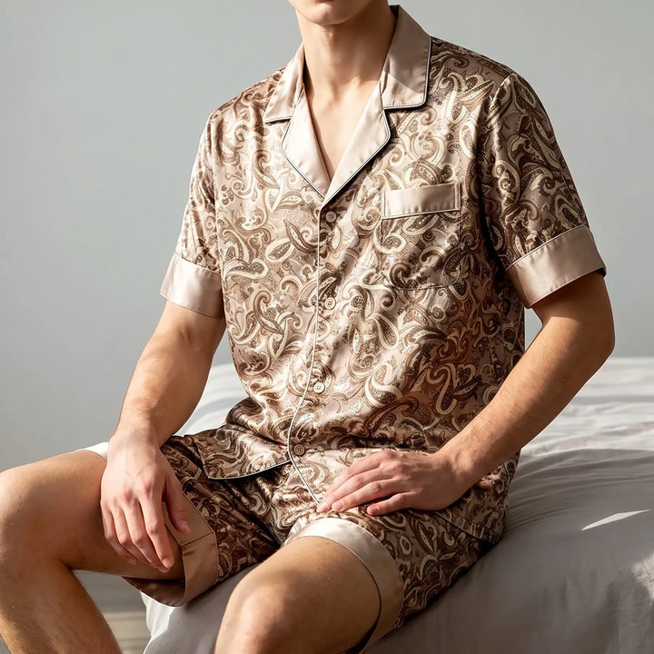 Model sitting on the edge of a bed in a satin paisley short-sleeve pajama set with tan contrast trim. 1