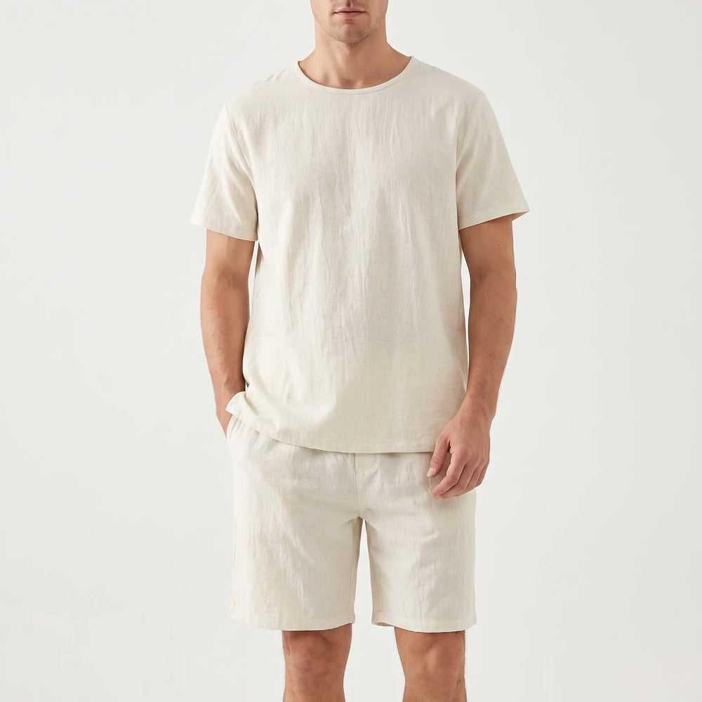 Front view on a plain backdrop of the cream pajama tee and shorts, one hand tucked into a shorts pocket. 2