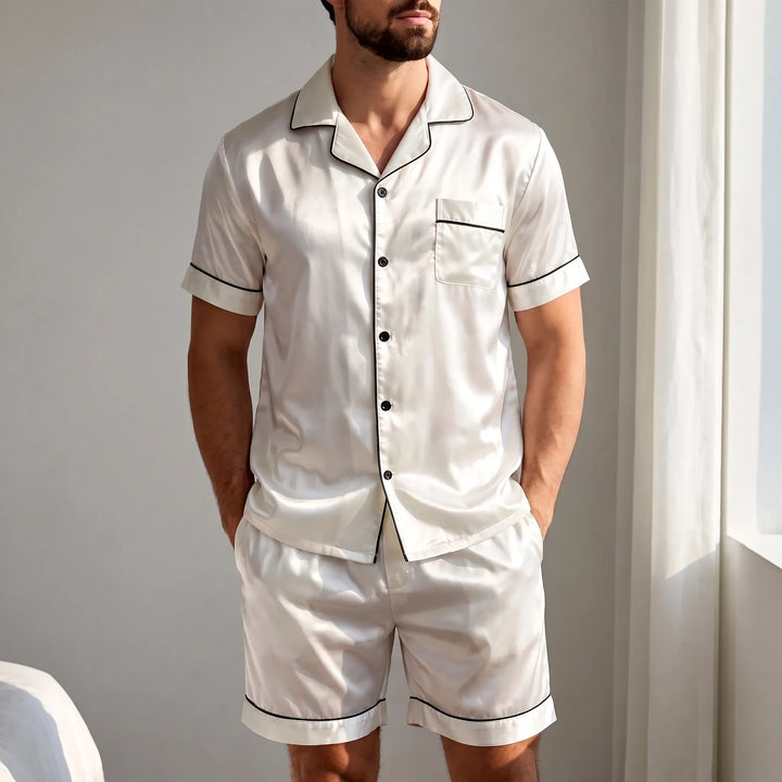 Bearded man in a sunlit bedroom wearing the white satin pajama set with black piping, hands in the shorts pockets. 1