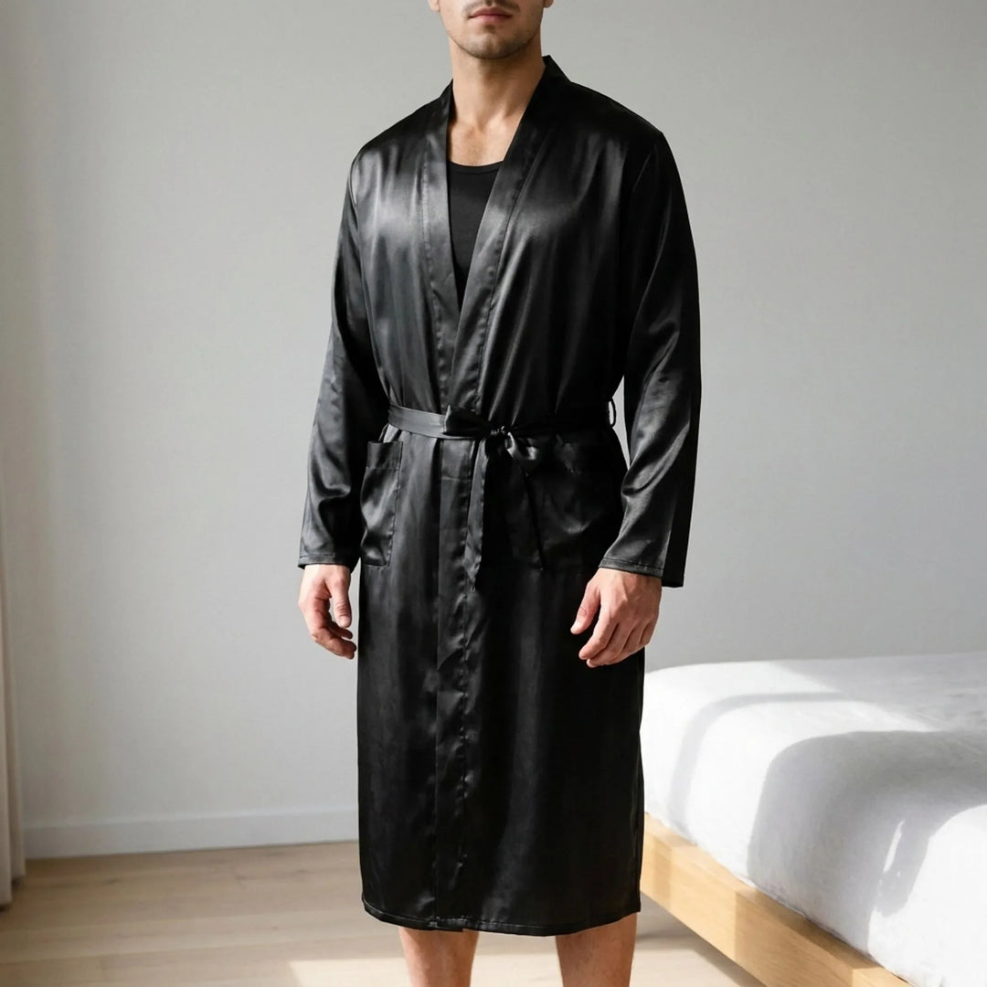 Man in a black satin robe tied at the waist over a dark tank top, standing in a bedroom beside a bed. 1