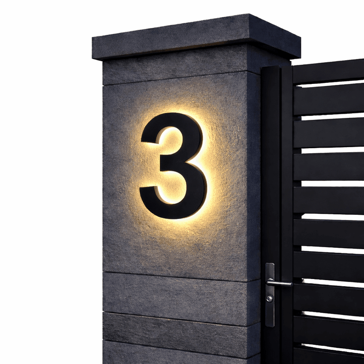Black backlit house number 3 with a warm glow on a dark stone gate pillar beside a black slatted gate.
