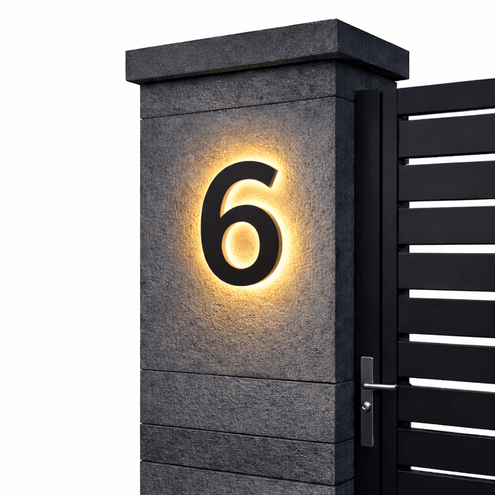 Black backlit house number 6 with a warm glow on a dark stone gate pillar beside a black slatted gate.