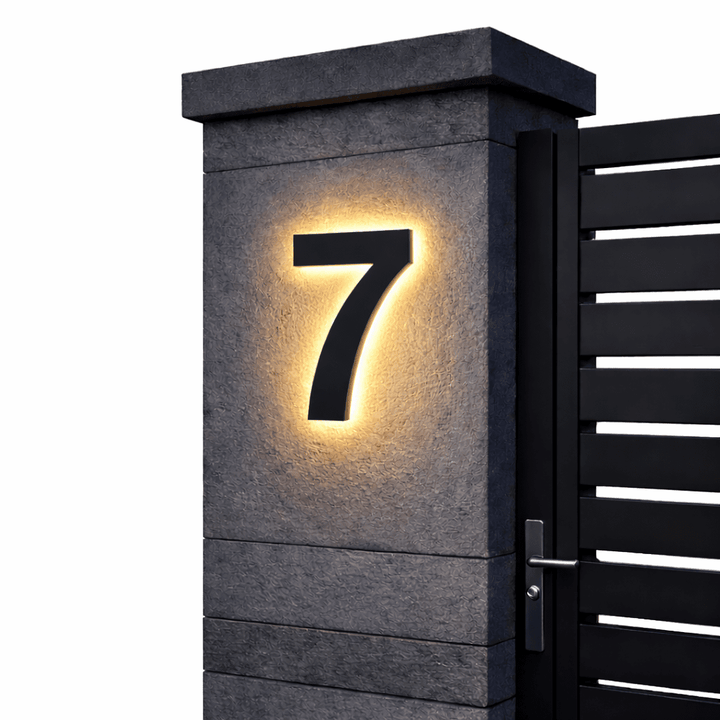 Black backlit house number 7 with a warm glow on a dark stone gate pillar beside a black slatted gate.