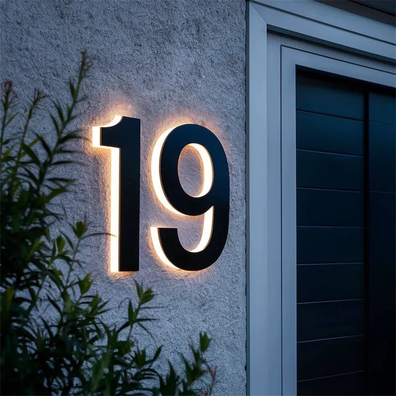 Close view of black two-digit house number with warm backlight on rough gray stucco.