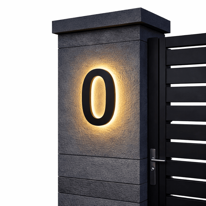 Black backlit house number 0 with a warm glow on a dark stone gate pillar beside a black slatted gate.
