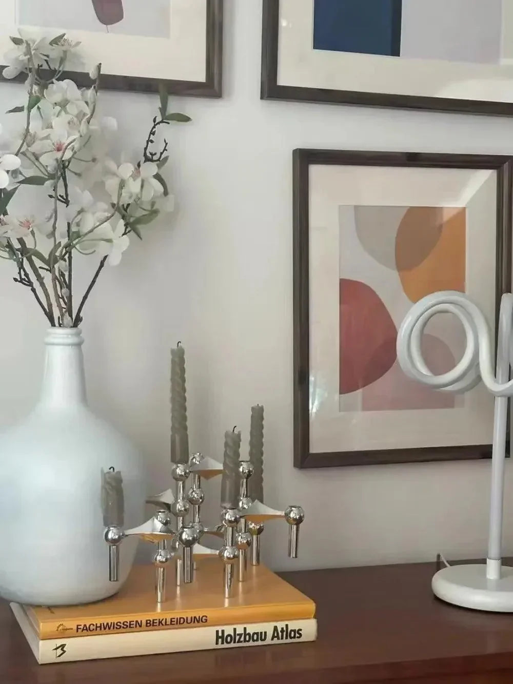 Silver modular candle holders on a stack of books beside a white vase of blossoms and framed art.