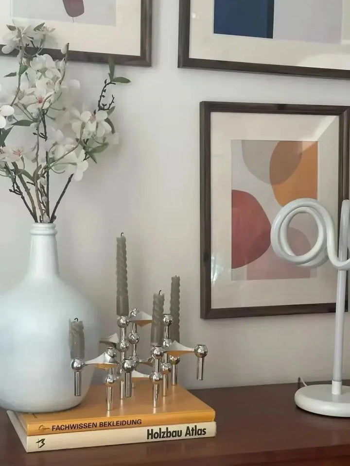 Silver modular candle holders on a stack of books beside a white vase of blossoms and framed art.