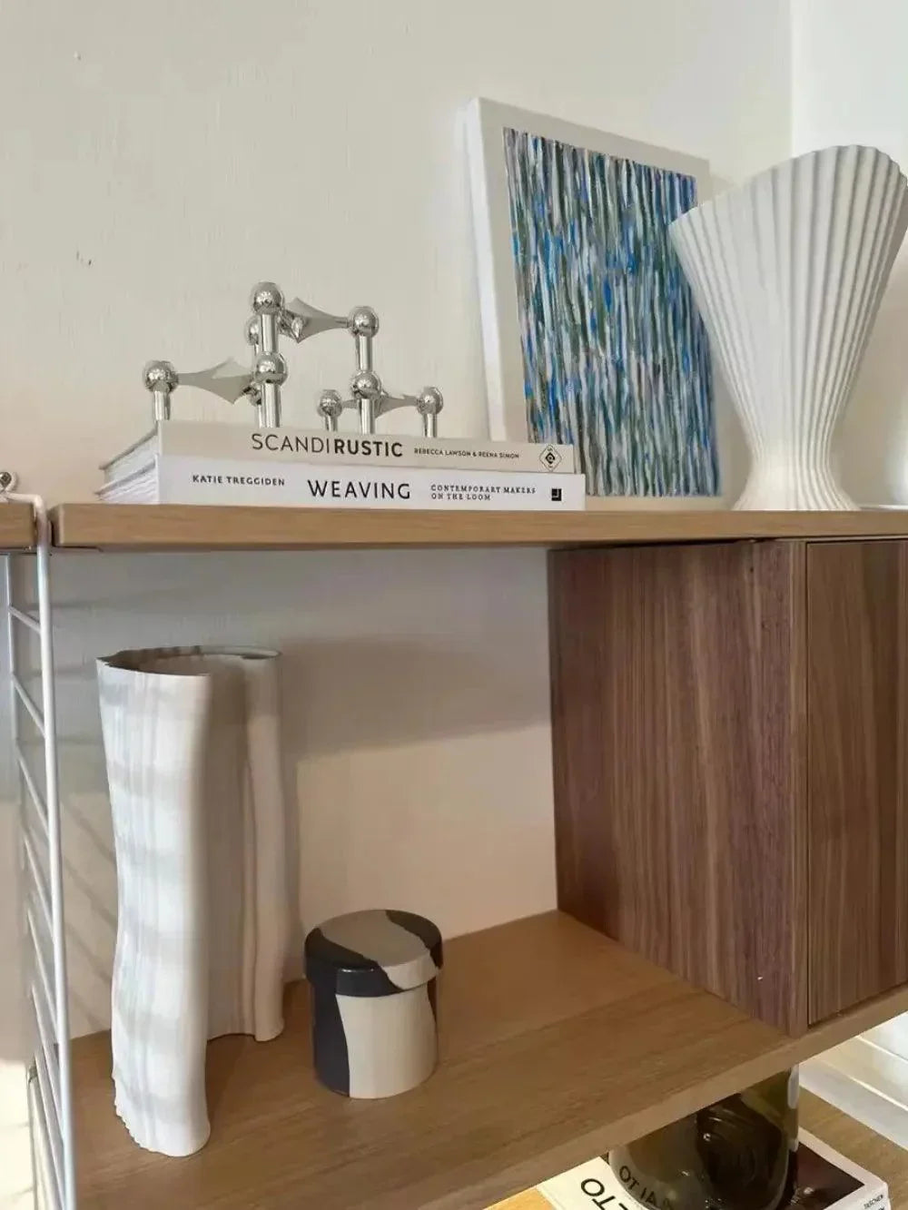 Silver modular candle holder arrangement on two books on a wooden shelf next to abstract artwork and white vases.