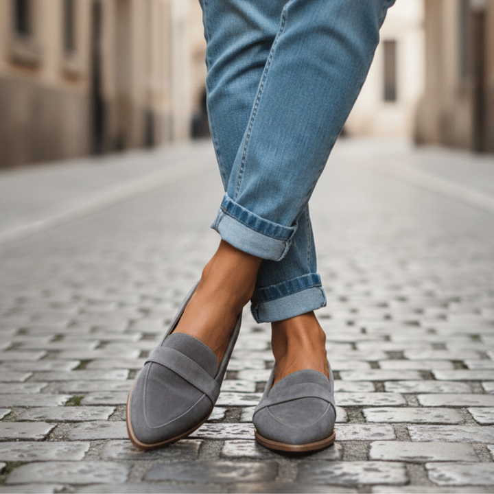 Woman wearing gray suede pointed-toe loafers with rolled light-wash jeans, legs crossed on a cobblestone street. 1