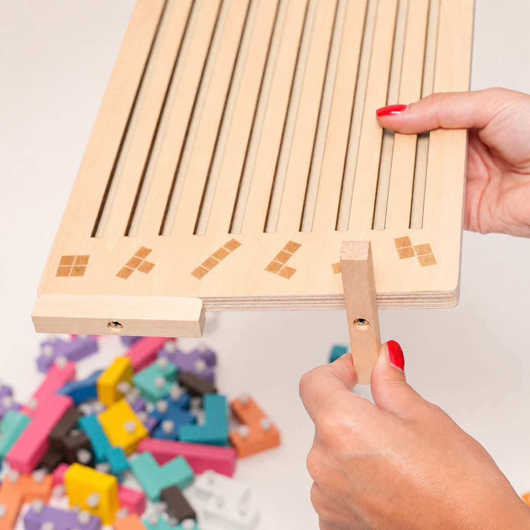 Hands attach a wooden support foot to the bottom edge of the slotted board, with loose pegged blocks piled below. 14