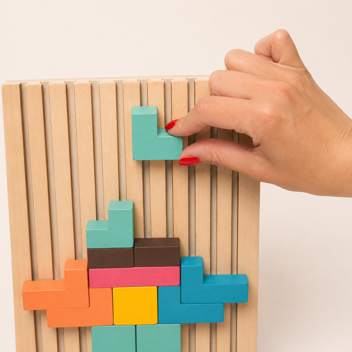Adult hand with red nails lowers a teal L-shaped block into the slotted board above a partly built colorful pattern. 12