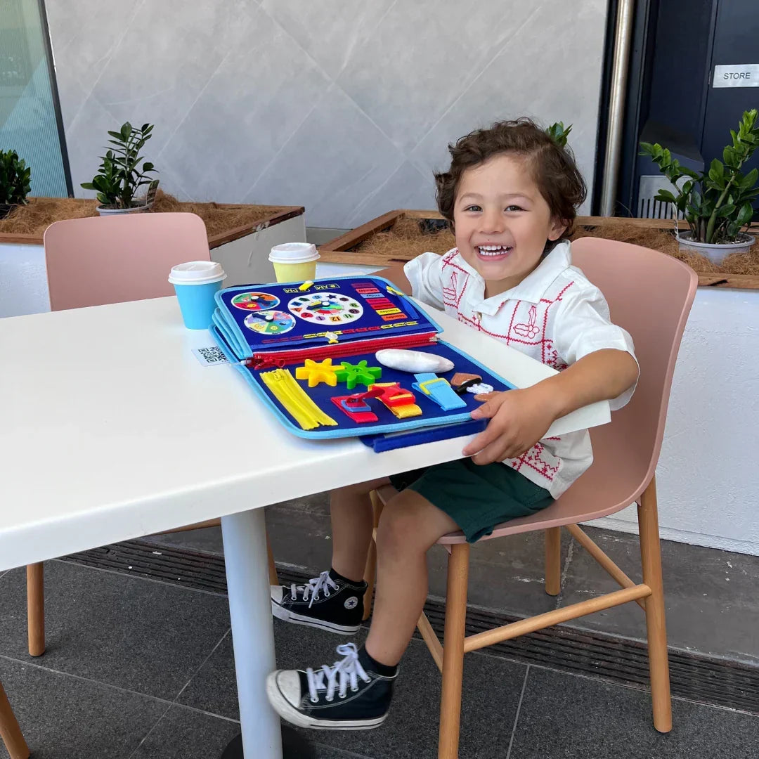 Laughing toddler boy at a white table with the blue felt activity book open, showing a clock page, gears and zippers. 7