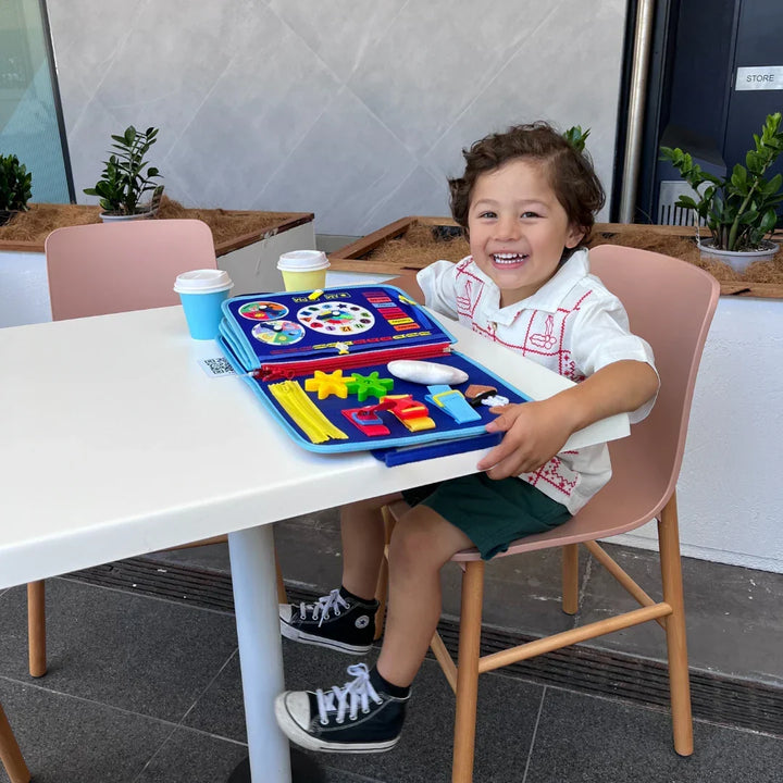 Laughing toddler boy at a white table with the blue felt activity book open, showing a clock page, gears and zippers. 7