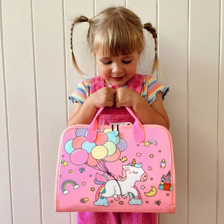 Girl with braided pigtails holding the closed pink carry case printed with a unicorn, balloons, rainbow and castle. 10