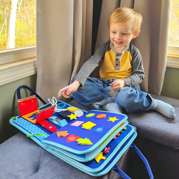 Smiling toddler boy sitting on a gray couch reaching for the blue felt activity book open on his lap, showing shapes and straps. 3