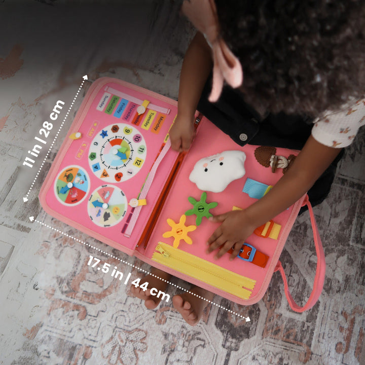 Overhead view of a child's hands on the open pink felt book, with dimensions marked across its clock, gear and zipper pages. 8