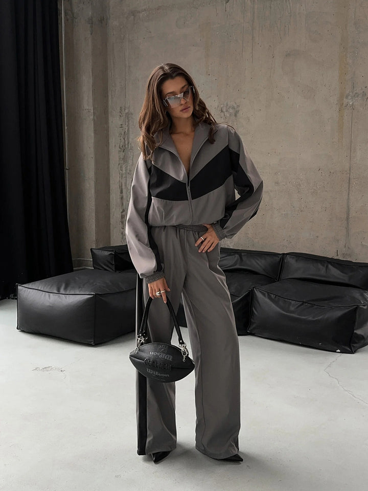 Model standing in the gray set with black chevron panels and zipped jacket, wide-leg pants, sunglasses, holding a black football-shaped handbag. 23
