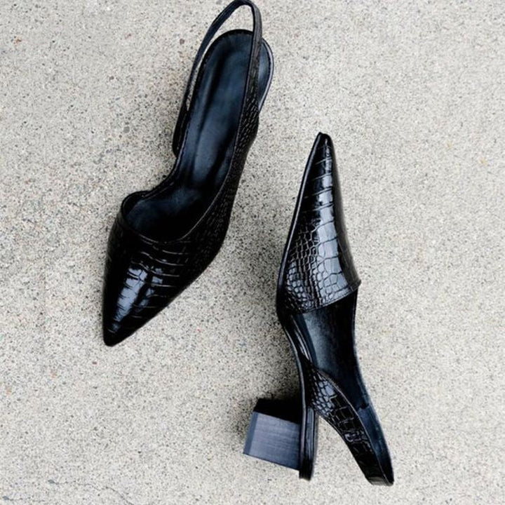 Flat lay of a pair of black croc-embossed slingback pumps with block heels on concrete, one shown from the top, the other on its side. 3