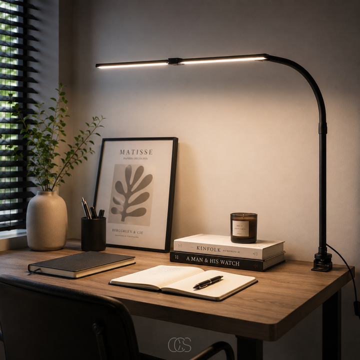 Black desk lamp another view with warm light over an open notebook, books, a candle, a framed print and a vase.