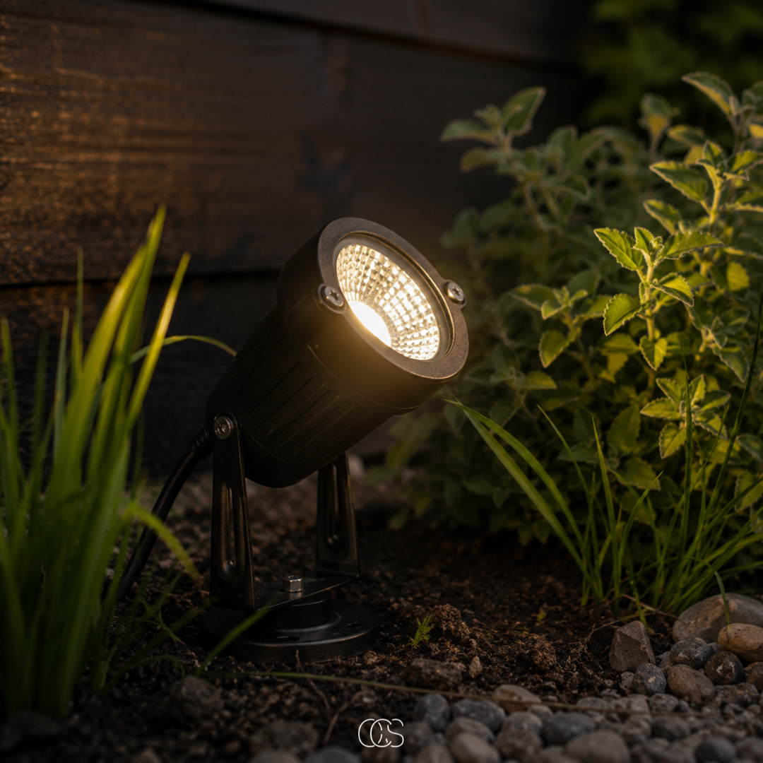 Black LED garden spotlight with ground stake and swivel head, glowing beside herbs and pebbles.