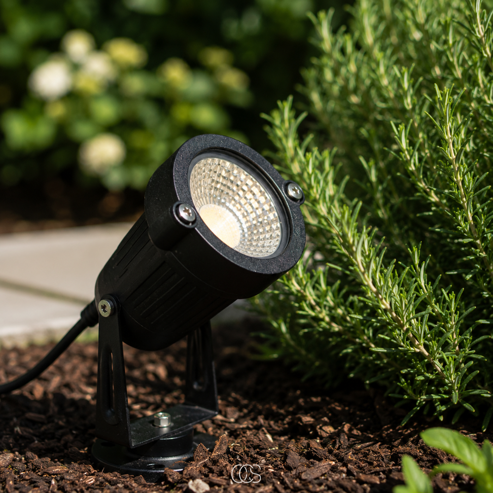 Black LED garden spotlight detail with screws and cable in bark mulch next to a rosemary bush.