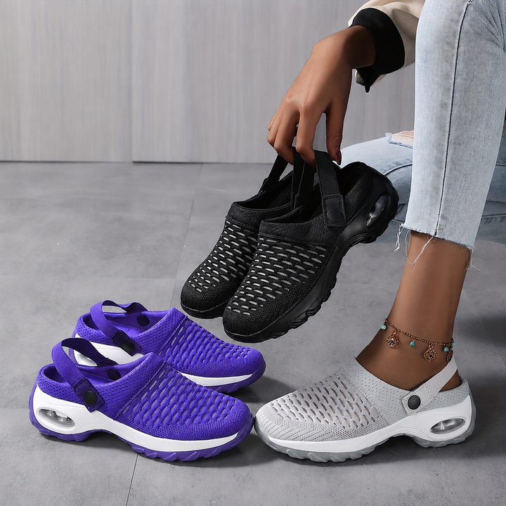 Three pairs of clogs in purple, black and gray on a concrete floor, one pair held up by a seated woman.