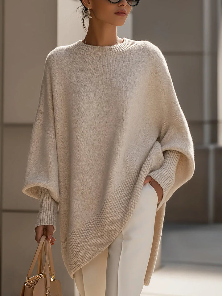 Model in a beige oversized knit sweater with ribbed cuffs and slanted hem, white pants, holding a tan bag 1