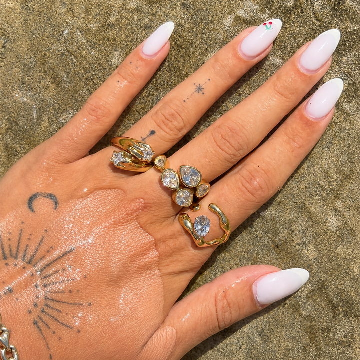 Hand with white manicured nails and small tattoos wearing the open gold ring with an oval clear stone, stacked with two other jeweled rings. 3