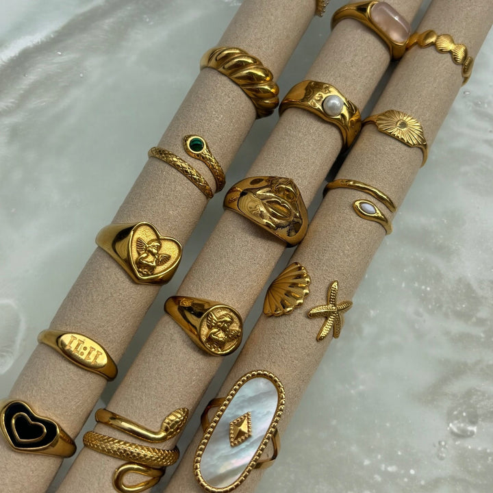 Flat lay of gold rings on beige tubes, including the heart cherub ring, snake, shell and pearl styles 5