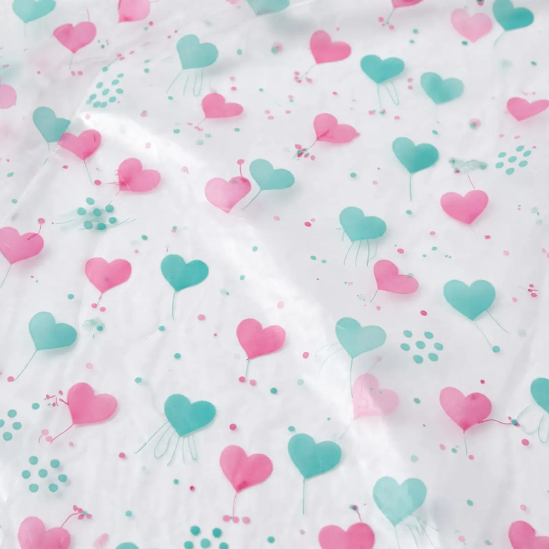 Close-up of the translucent raincoat material printed with pink and teal hearts and small dots, with soft creases catching the light. 6