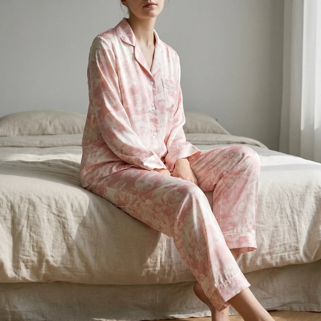 Woman sitting on the edge of a bed in the pink-and-white animal-scene pajama set with piped notch collar, in a sunlit bedroom. 1