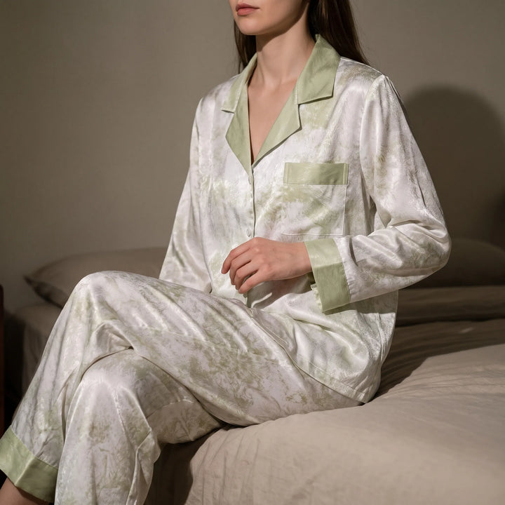 Model sitting on the edge of a bed in the satin-look pajamas, white with a soft green marbled print and green trim. 1