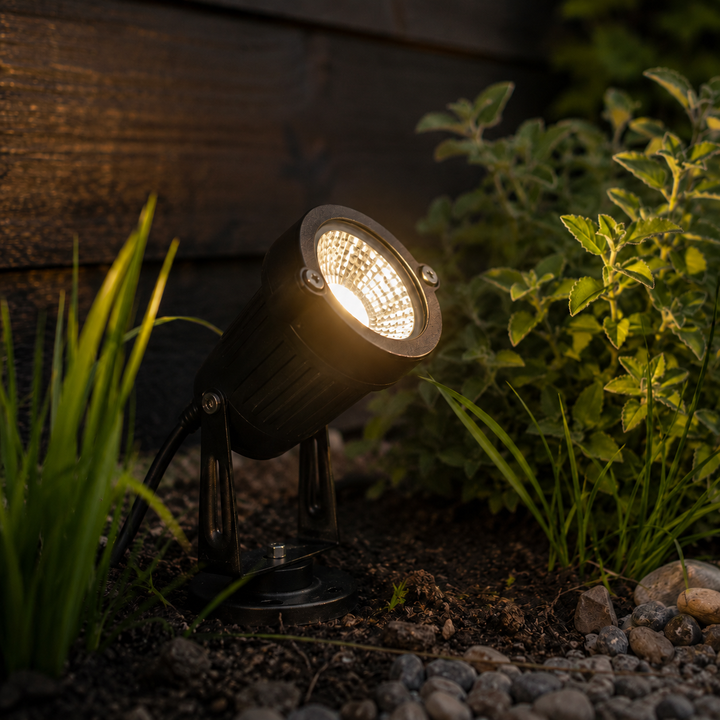 Black LED garden spotlight with ground base glowing in dark soil near pebbles and mint leaves.