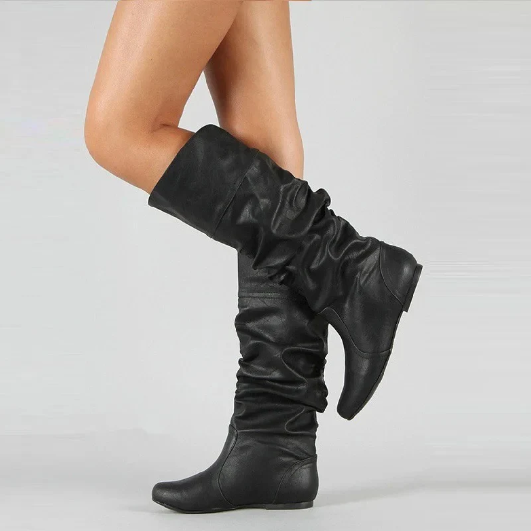 Side view of a model wearing black slouchy leather boots with a folded cuff and rounded toe, one leg bent.