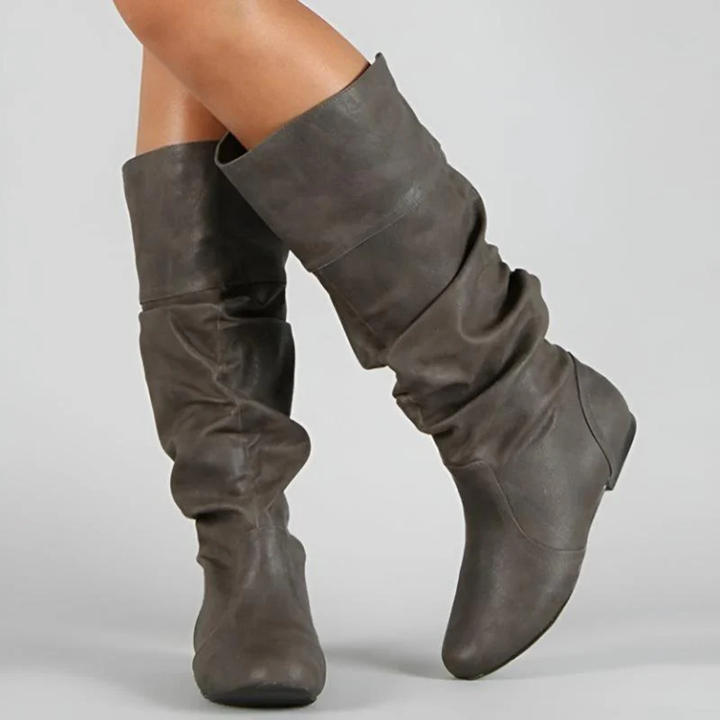 Close-up of a model wearing gray-brown slouchy mid-calf boots with a folded cuff, rounded toes and flat soles.