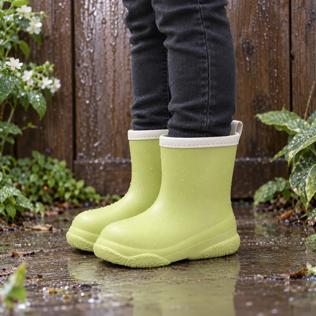 Child in dark jeans wearing light green rain boots with cream collars, standing in a rainy puddle by a wooden fence. 3