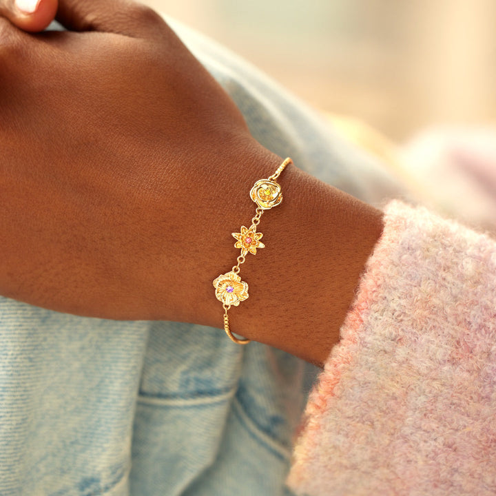 Gold birth flower bracelet worn on a wrist with a pink knit sleeve and denim jacket nearby.