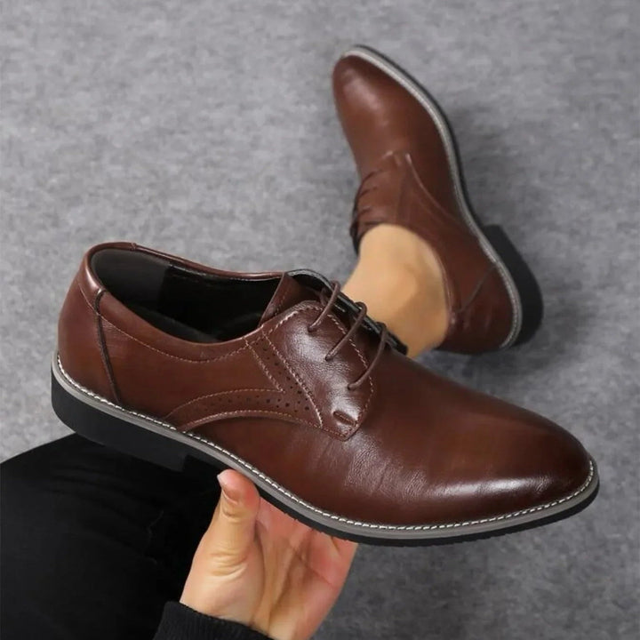 Dark brown polished lace-up shoe held in a hand above a gray carpet, with the second shoe behind it.