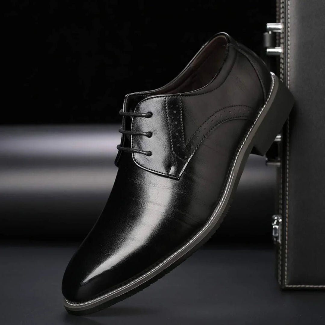 Single black polished lace-up shoe with perforated brogue detail and contrast stitching, shown leaning against a dark briefcase.