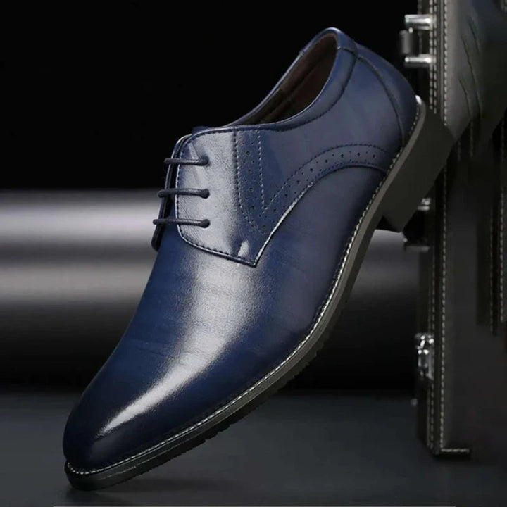 Single navy blue polished lace-up shoe with perforated brogue detail, shown against a dark background and briefcase.