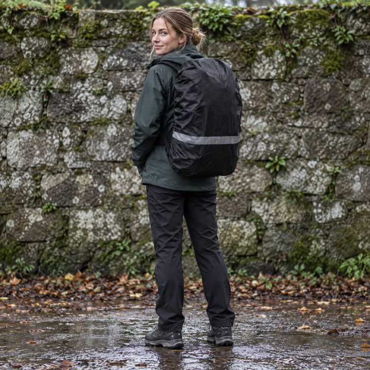 Woman in a dark green jacket looks over her shoulder wearing a rain-speckled black rucksack cover with a reflective stripe, standing before a mossy stone wall. 1