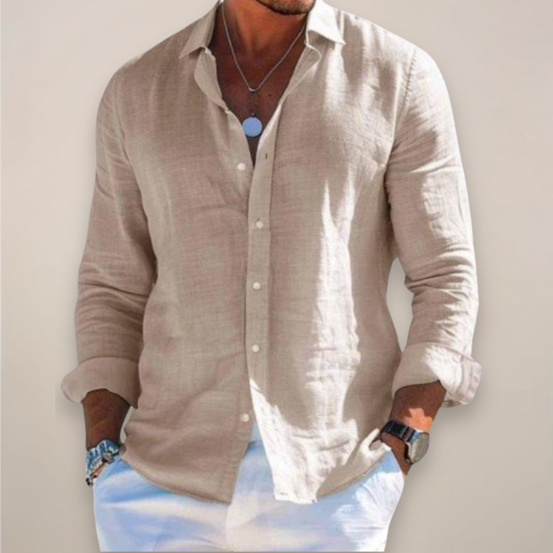 Man wearing the beige long-sleeved button-front shirt with rolled sleeves, hands in white pants pockets and a pendant necklace.