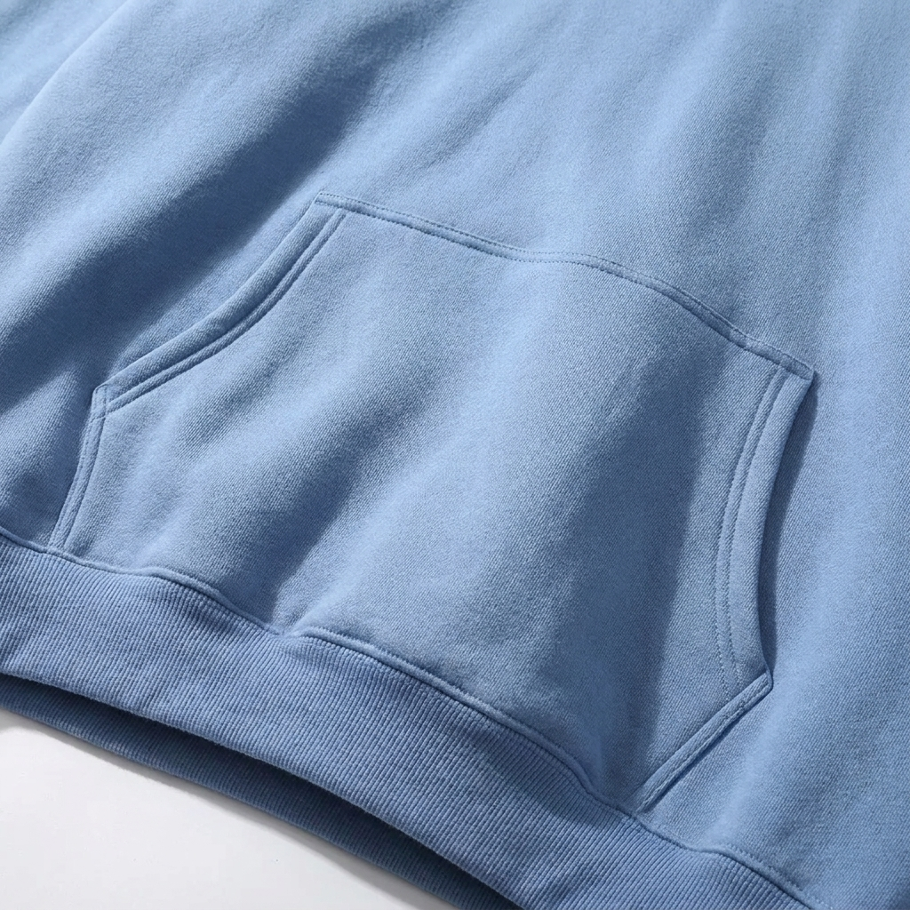 Close-up of the light blue kangaroo pocket, showing its stitched edges, soft fleece texture and the ribbed hem below.