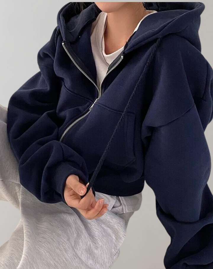 Navy blue zip-up hoodie worn open over a white top and gray pants, with a hand holding the drawstring.