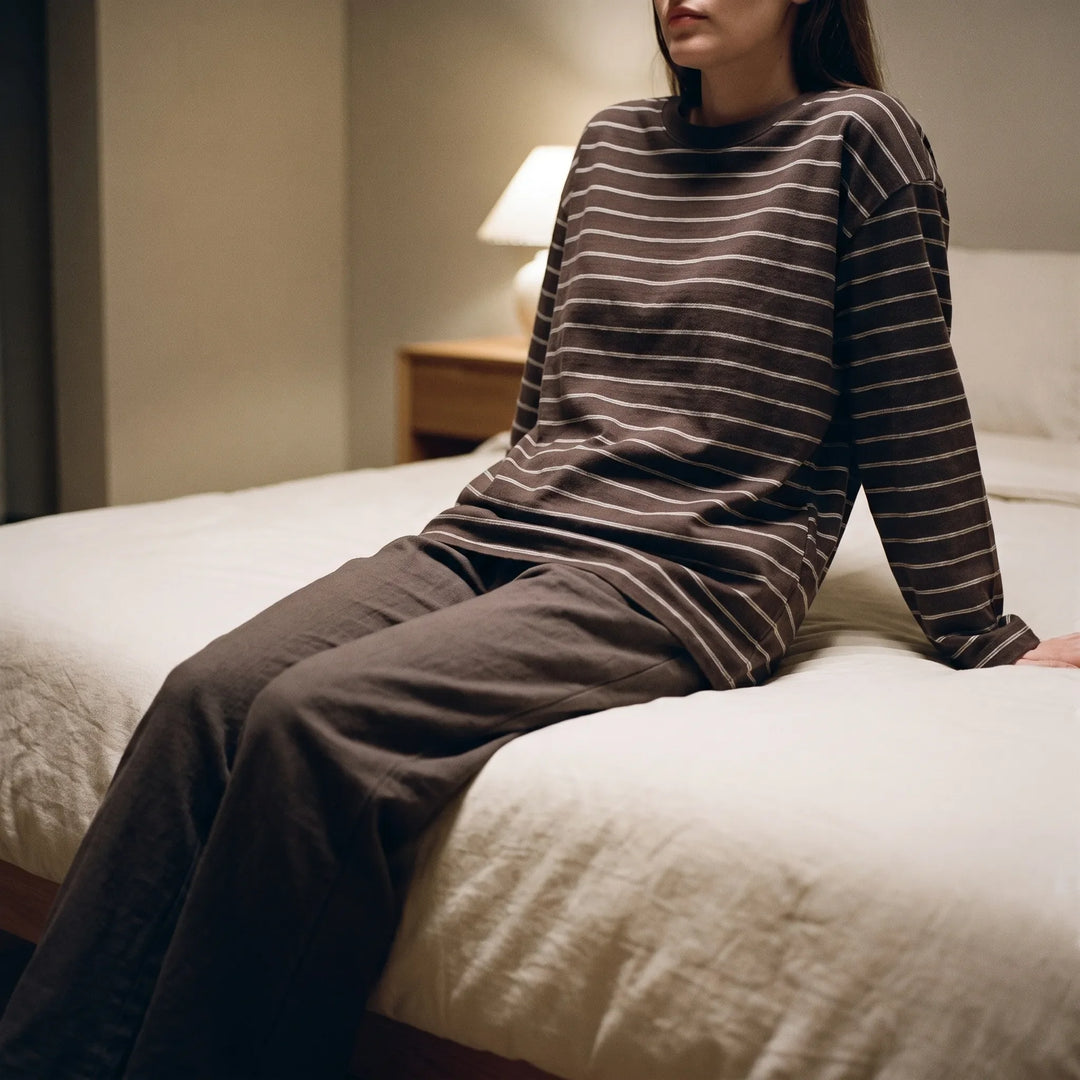 Model sitting on the edge of a bed in a dim bedroom wearing the brown striped long-sleeve top and matching loose brown pants. 1