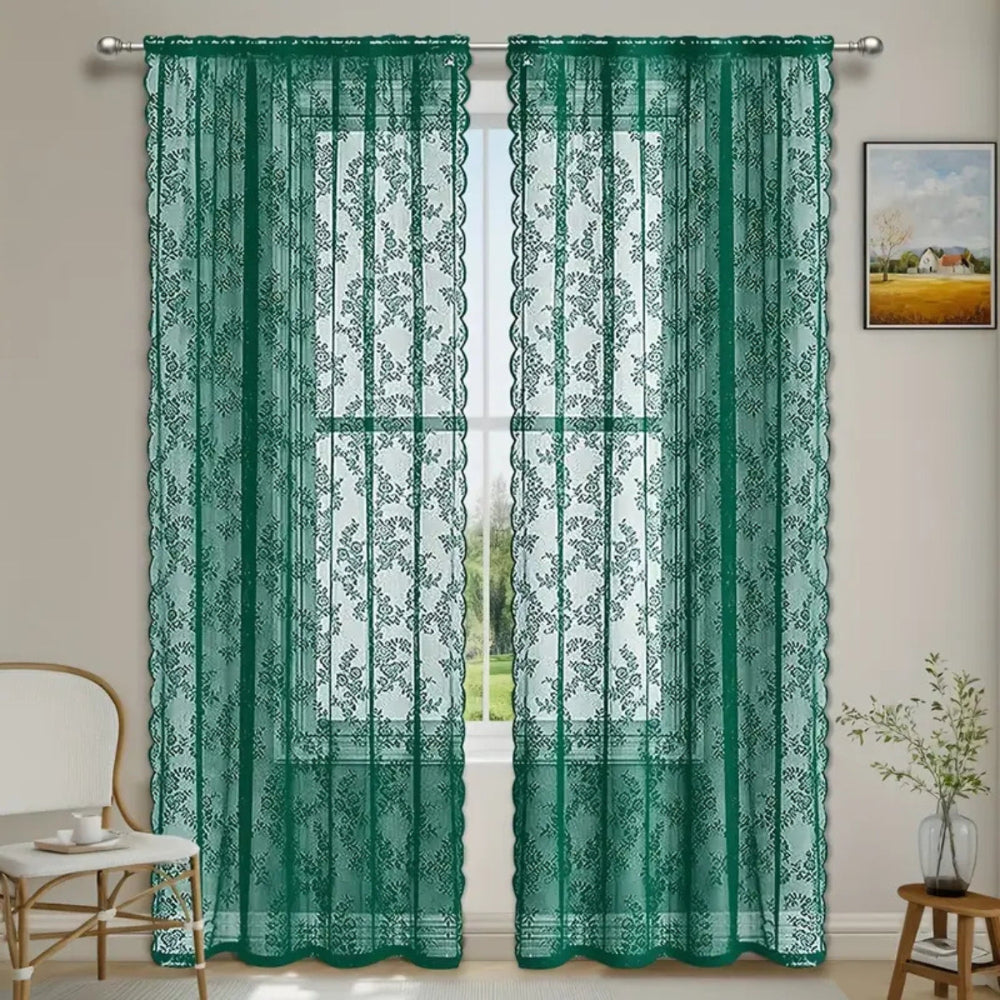 Pair of dark green floral lace panels hanging from a silver rod at a bright window, next to a chair and plant.