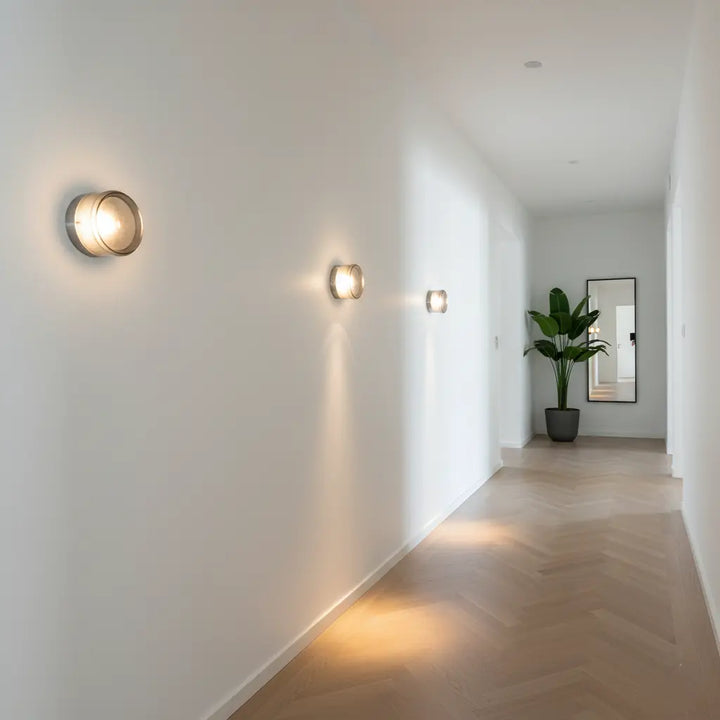 Three round glass wall lights glowing on a white hallway wall with a herringbone wood floor.