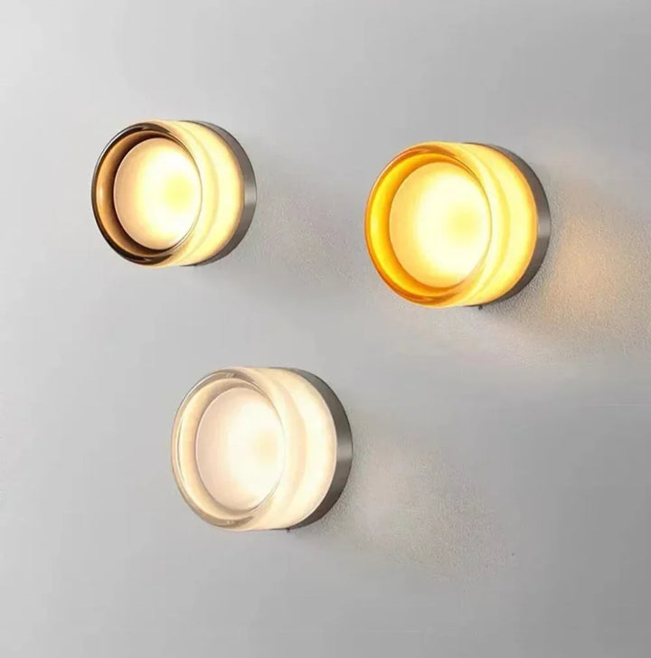 Three lit round glass wall lights in smoky gray, amber and clear glass on a light gray wall.