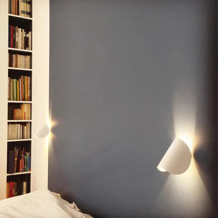 Two white wall lights tilted on a blue-gray bedroom wall next to a tall bookshelf, glowing softly.